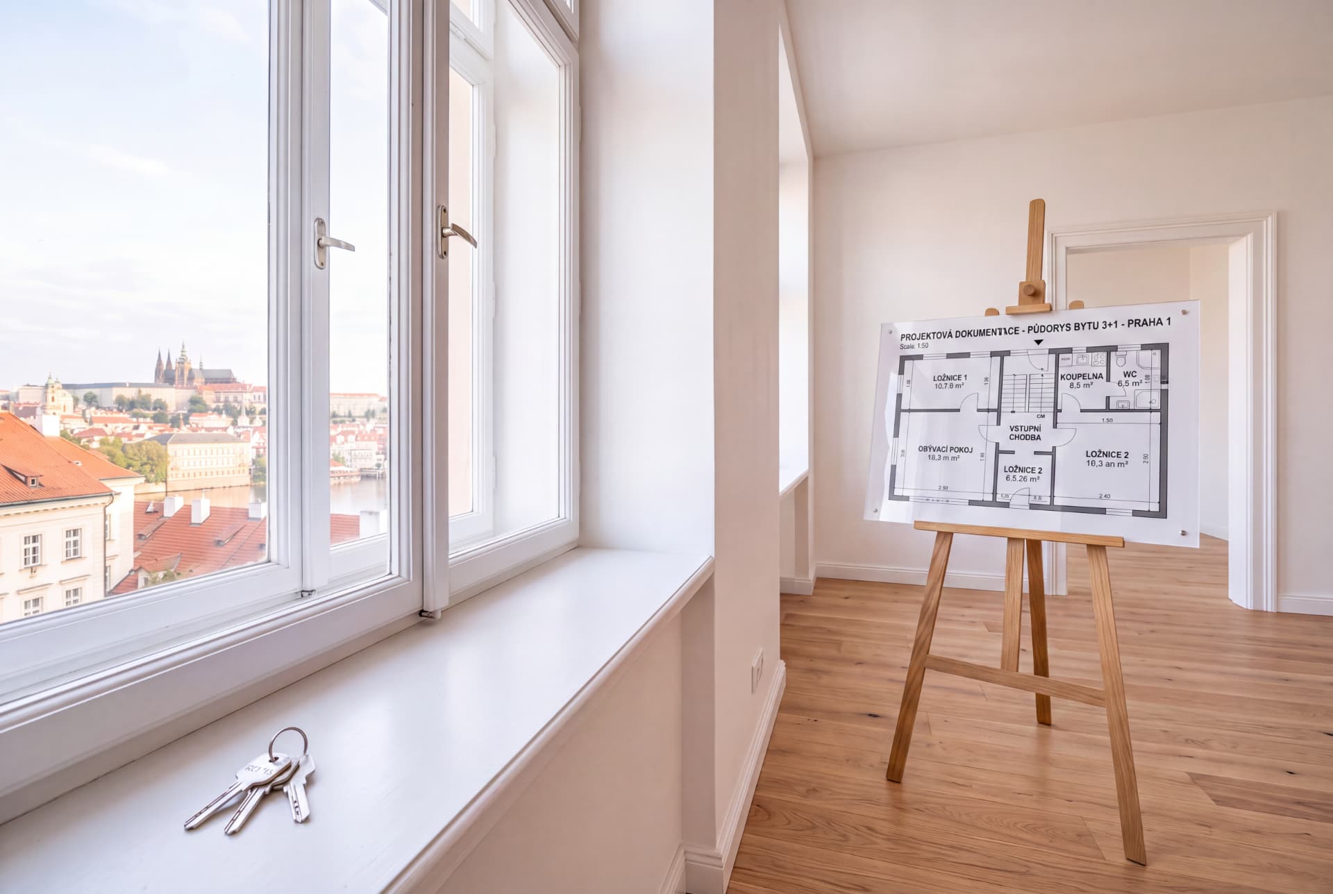 Prázdný byt s dřevěnou podlahou, velkými okny a výhledem na pražské střechy — na stojanu projekový půdorys bytu 3+1 připraveného k pronájmu